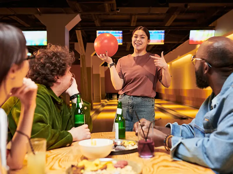 Coworker holding up a bowling ball while chatting with colleagues at a table during a team-building event at Jax 60 in Jackson, Michigan